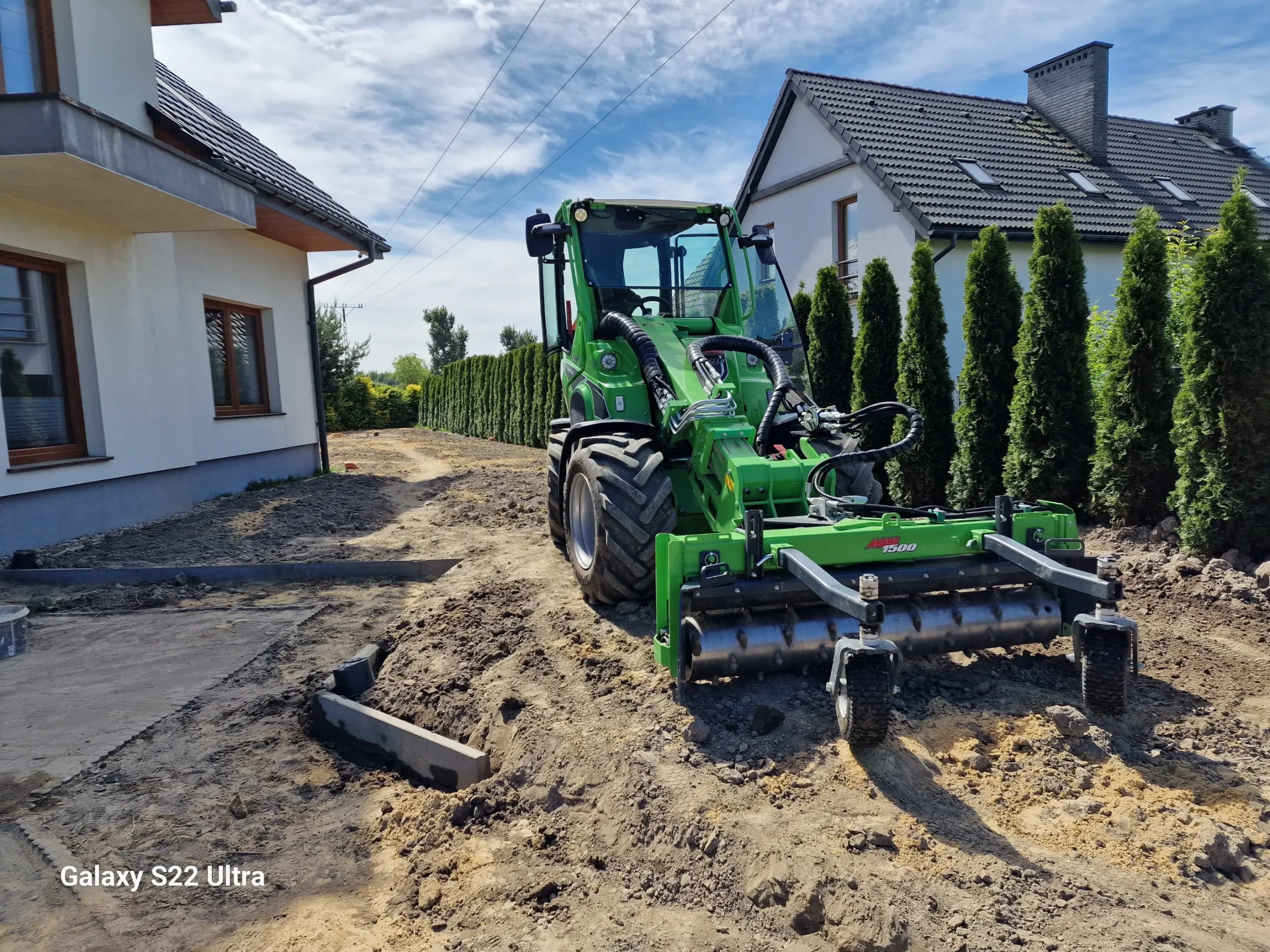 Praca glebogryzarką: zielona maszyna wyrównuje i spulchnia glebę przed domem przy żywopłocie
